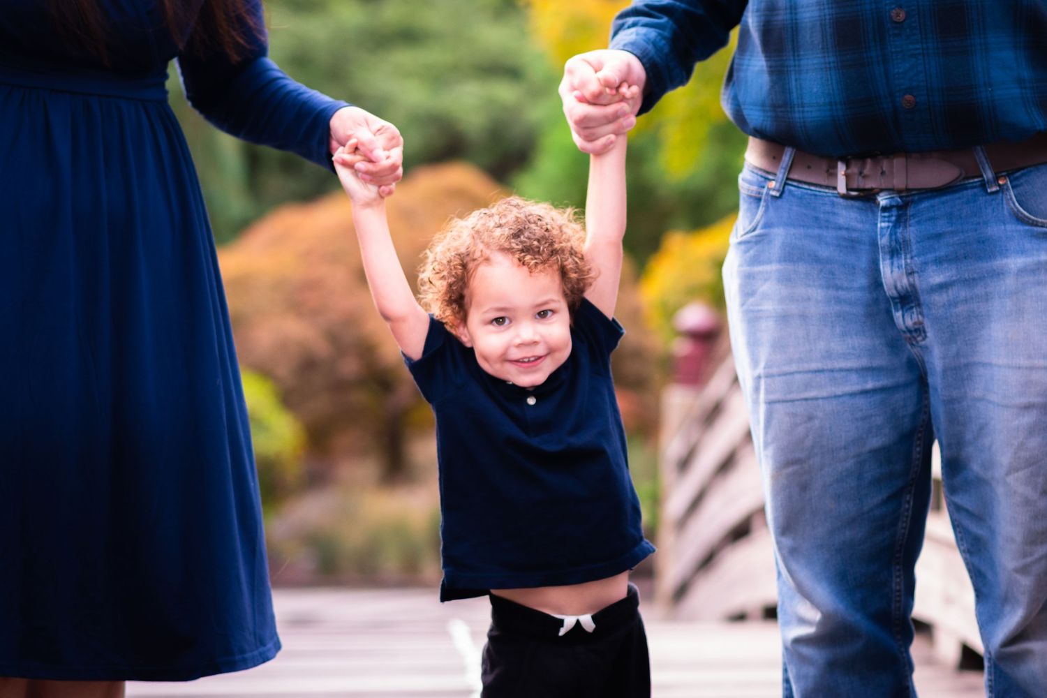 A happy toddler with curly hair holds hands with parents outdoors, smiling joyfully on a wooden bridge.