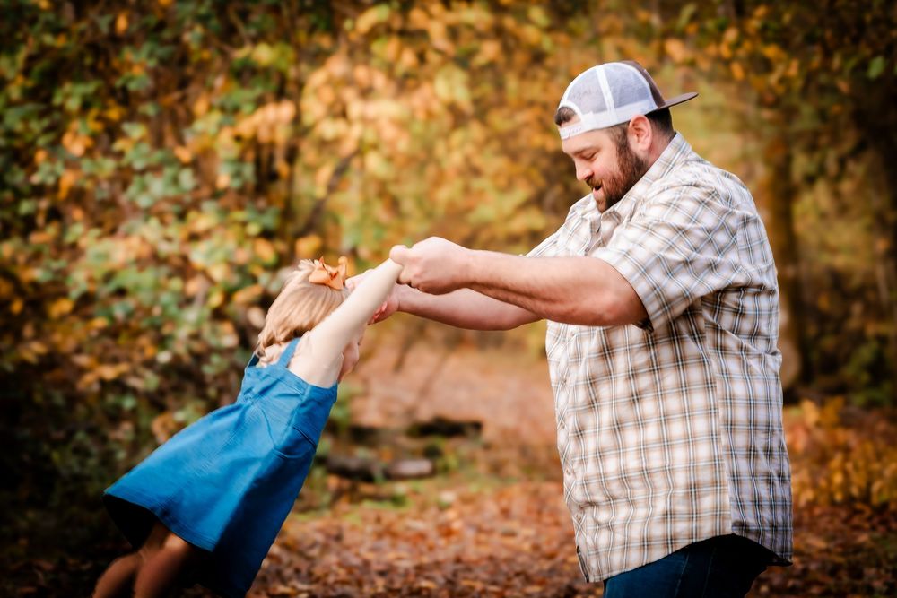 A father and young child hold hands and dance together in an autumn forest setting.