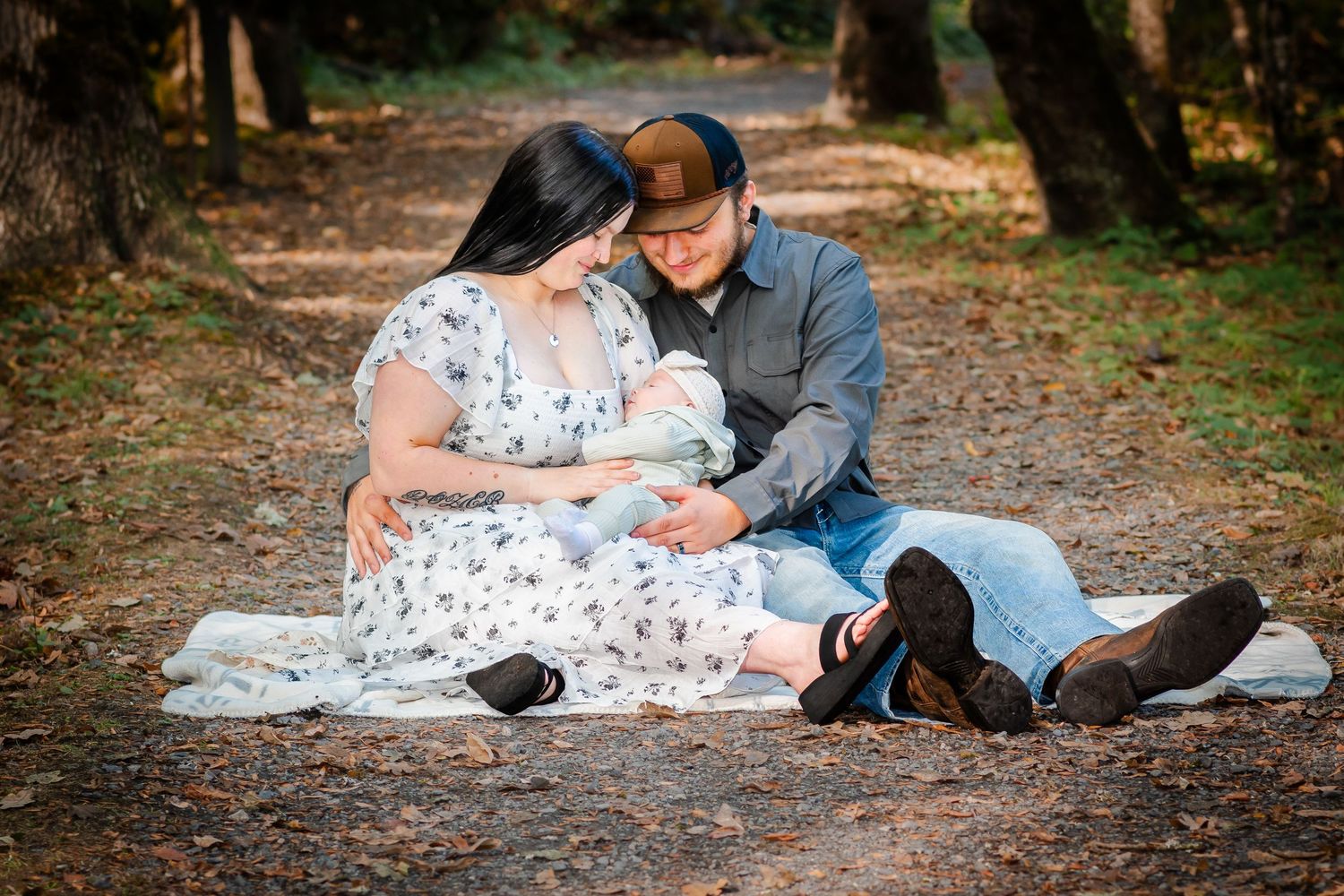 A couple sits on a blanket in a forest path, lovingly gazing at their newborn baby during an outdoor family photo session.
