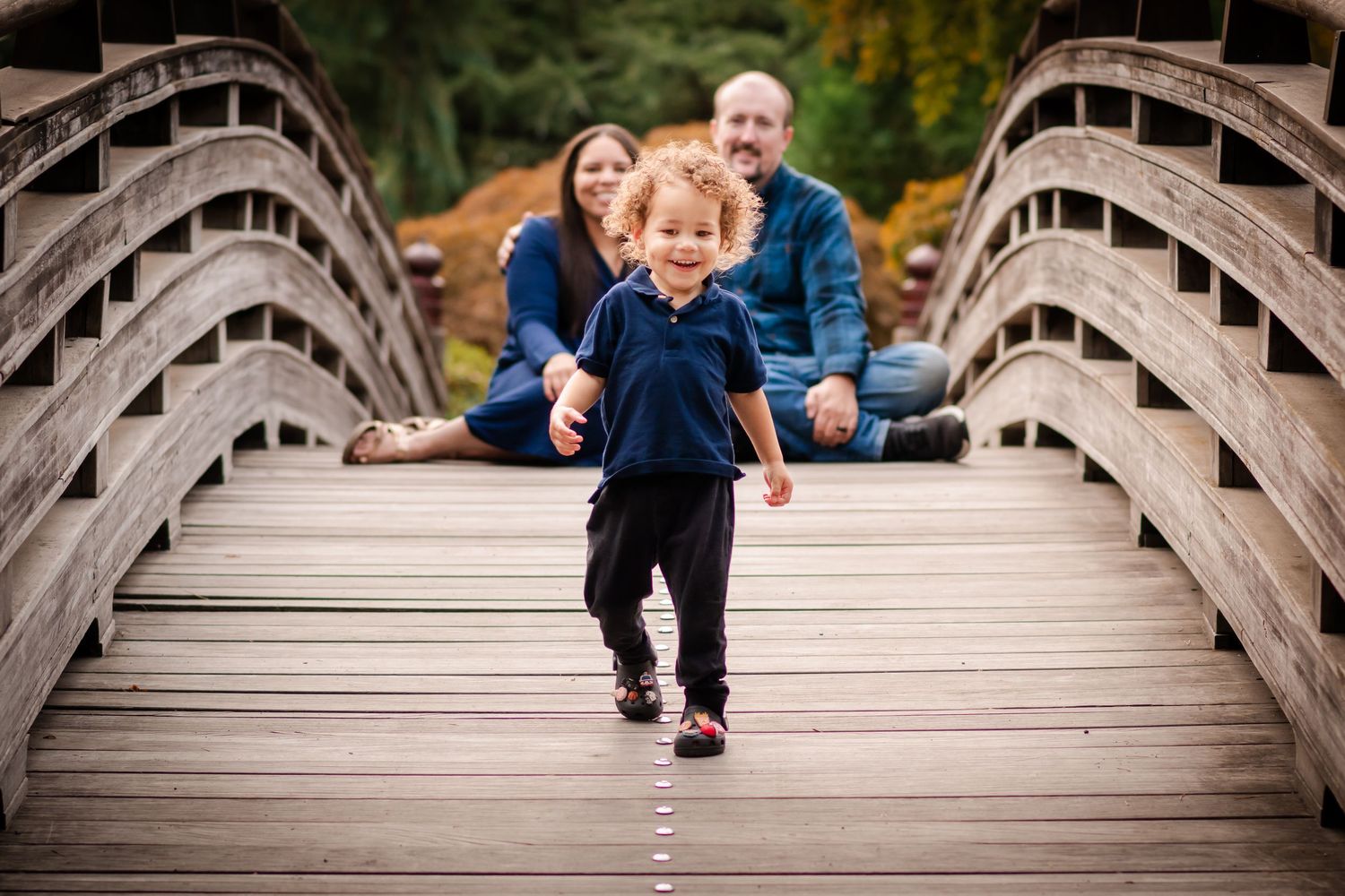 A young toddler runs on a wooden bridge while his parents sit smiling in the background during a family photo session.