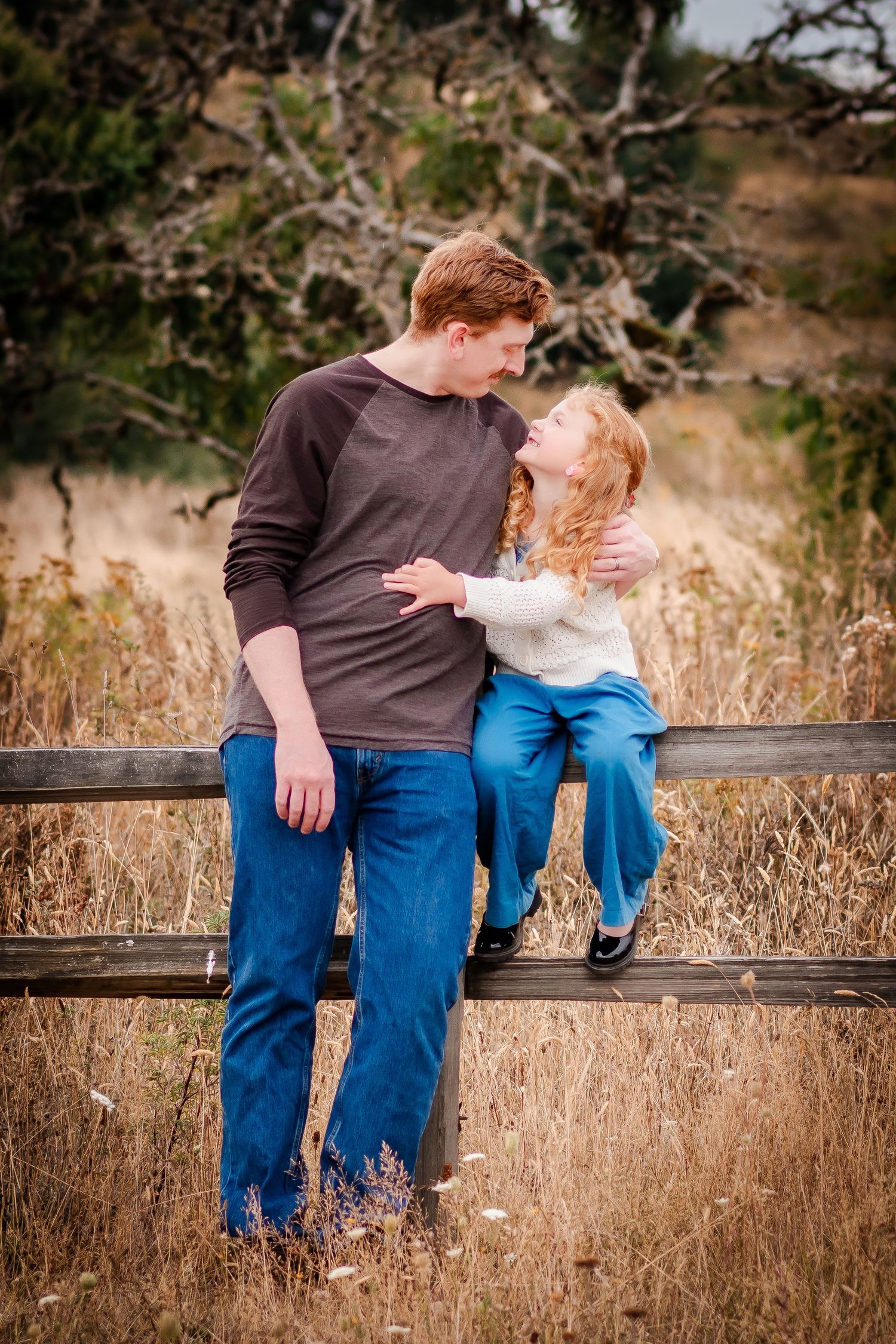 Couple sitting together on wooden fence surrounded by golden dried grass in autumn nature setting.