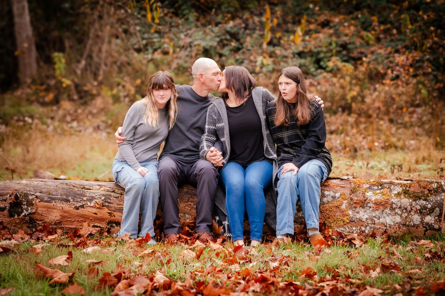 Family of four sitting on a log surrounded by autumn leaves in a wooded setting during fall season.