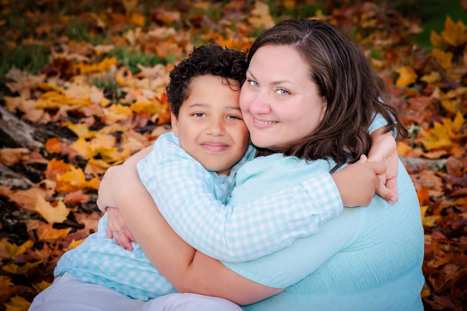 A mother and son embrace warmly among colorful autumn leaves, both wearing light blue, smiling in an outdoor fall setting.