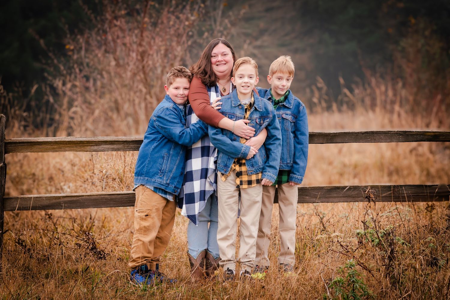 A woman and three boys in denim jackets pose together by a wooden fence in an autumn field.