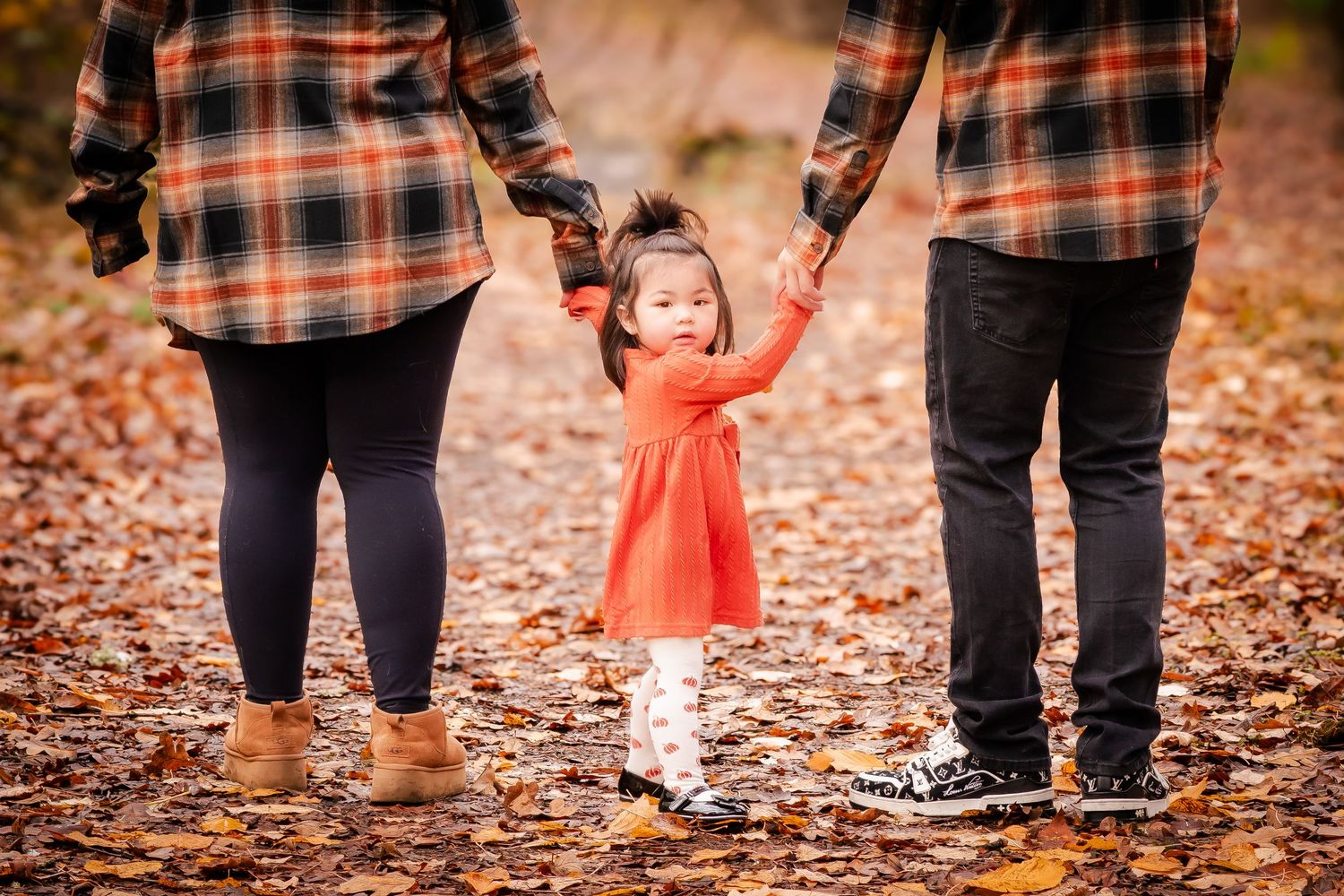 Toddler girl in red dress holds parents hands on autumn leaf-covered path during fall family photo session.