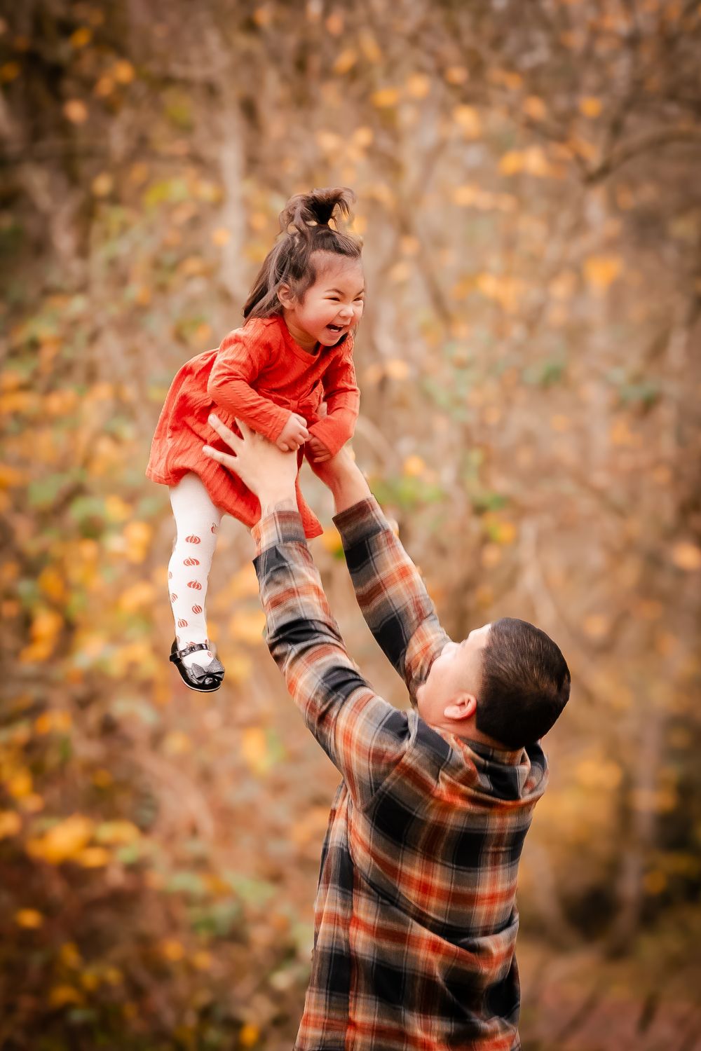 Father tossing laughing little girl in red dress into the air during autumn outdoor family photo session.
