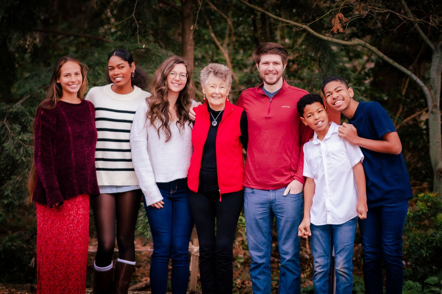 A multigenerational family of seven smiling together outdoors in front of lush green trees in a warm group portrait.