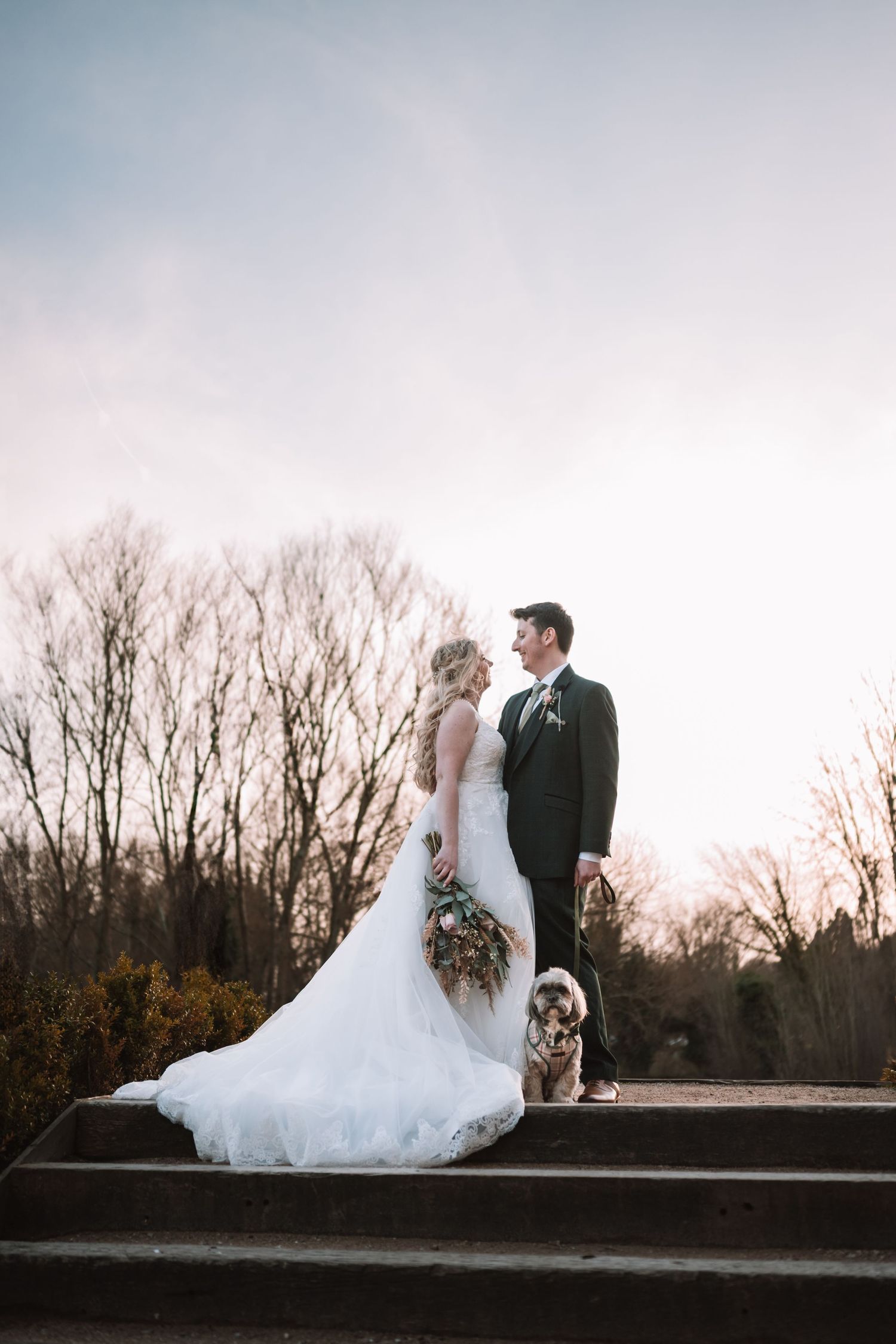 A romantic wedding couple stands on stone steps at sunset with bare winter trees in the background at the West Mill, Derby.