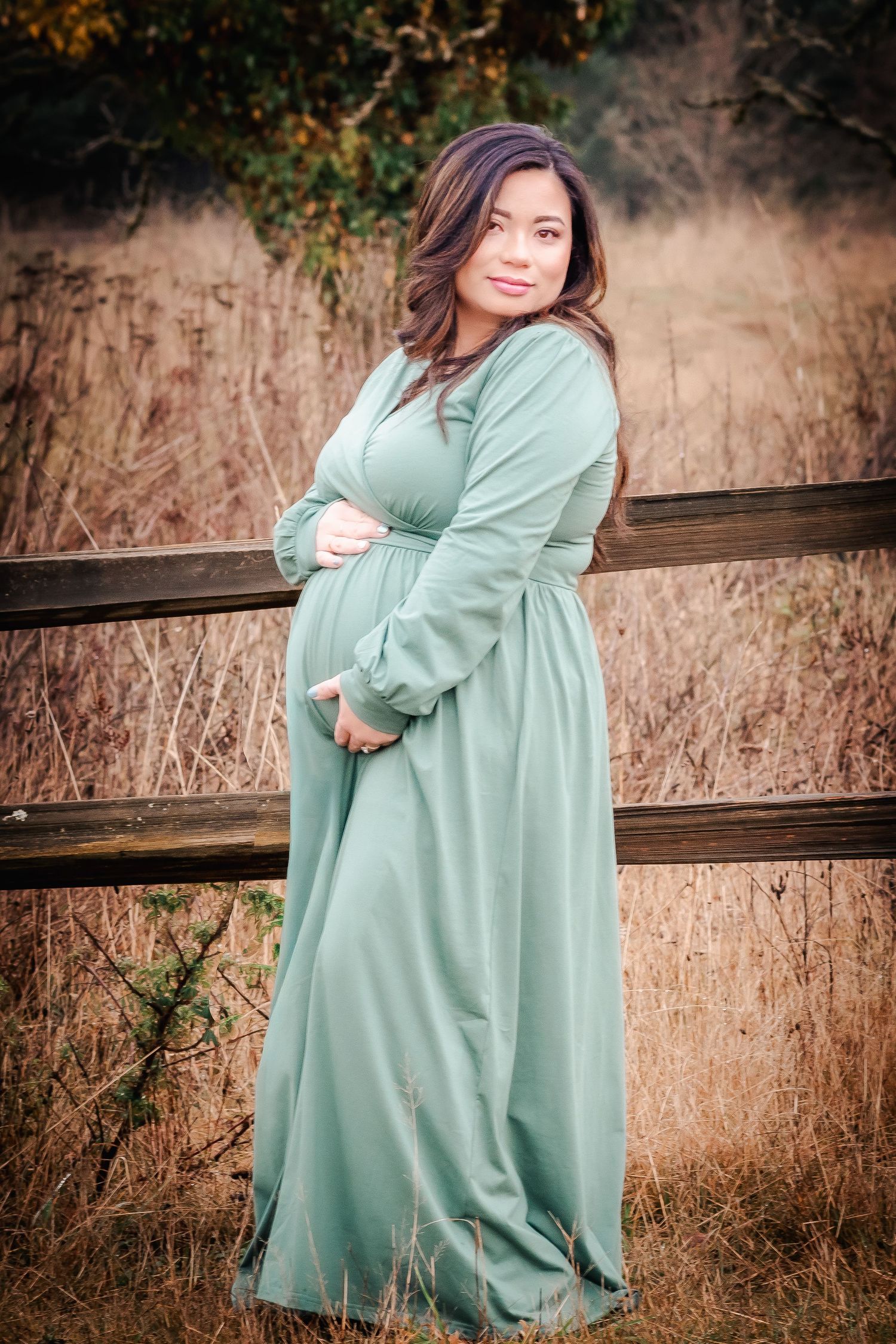 Pregnant woman in sage green maxi dress poses by wooden fence in field during maternity photo session.