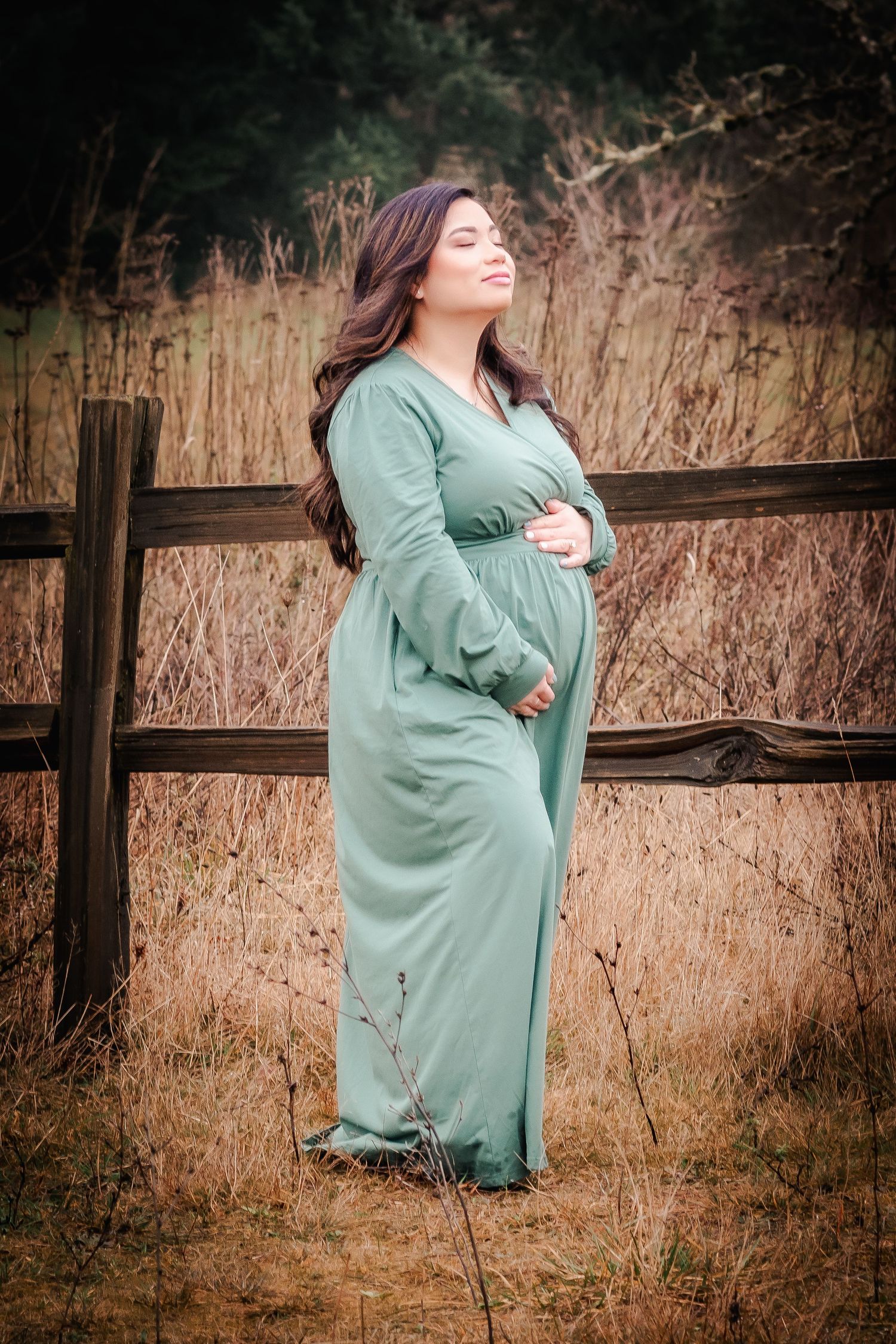 Pregnant woman in sage green maxi dress poses by rustic fence in golden field.