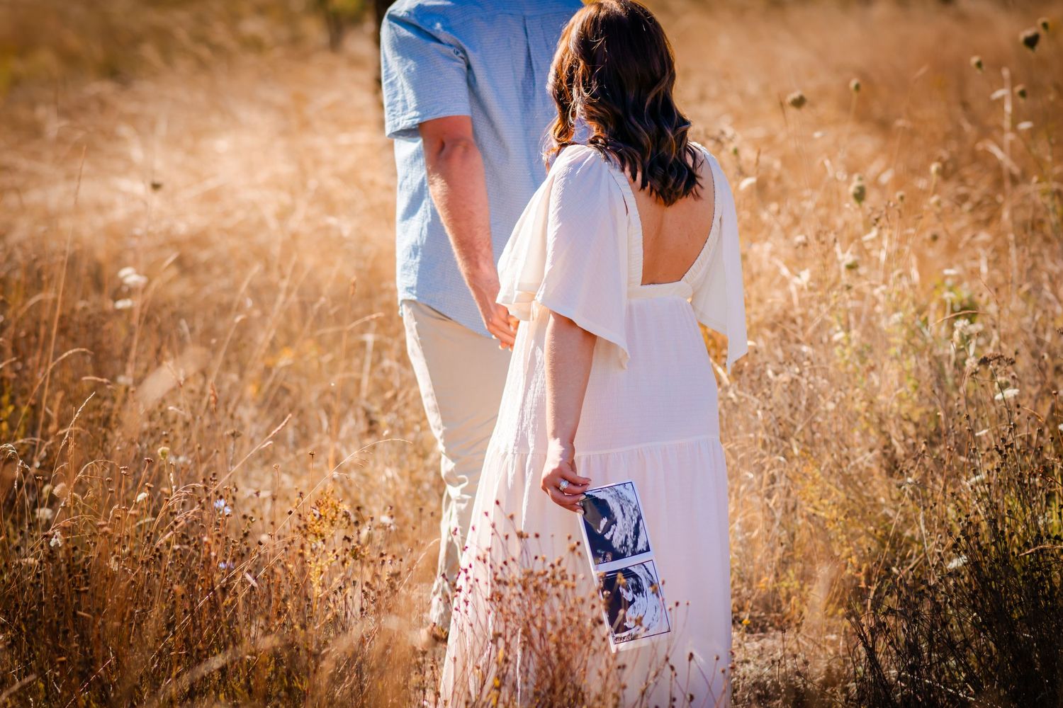A couple in white outfits poses intimately in a golden sunlit field.