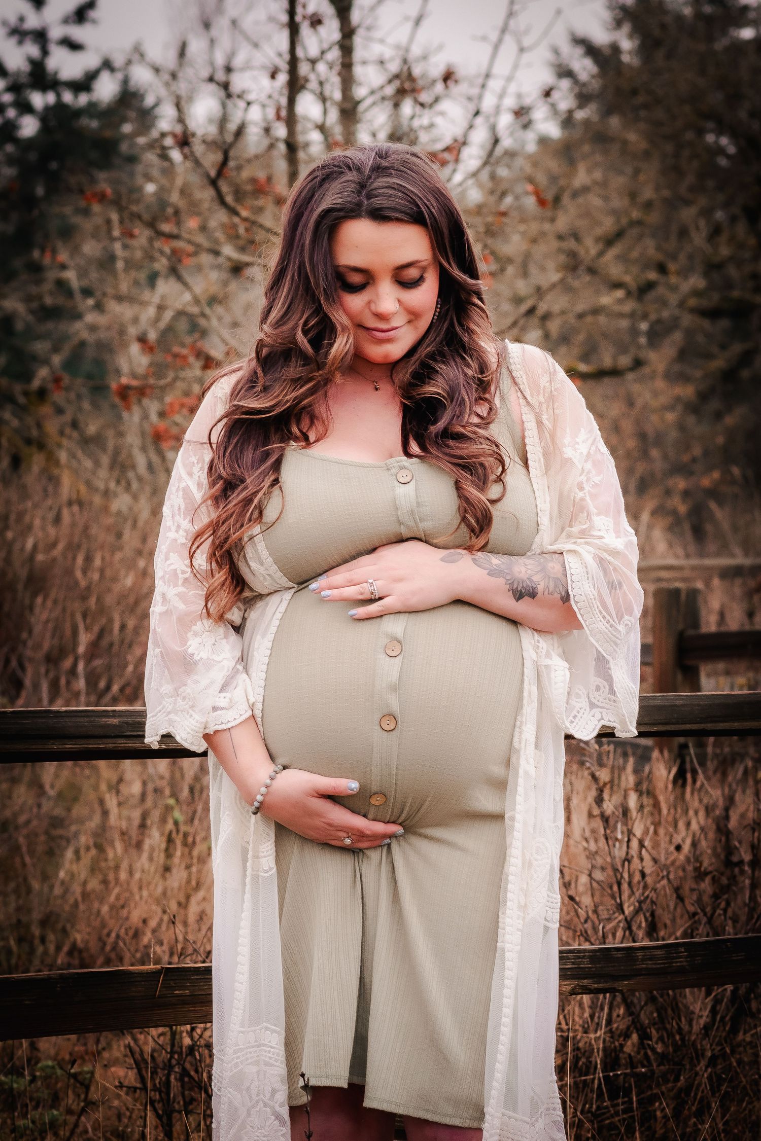 Pregnant woman in sage green dress and lace kimono cradles baby bump outdoors near wooden fence.