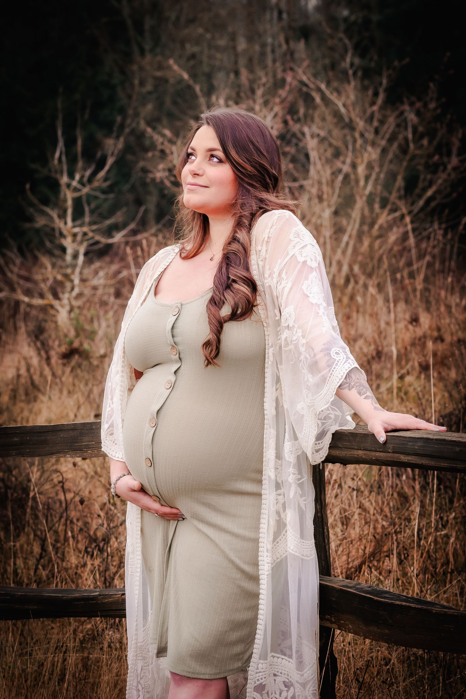 Pregnant woman in sage green dress and lace kimono cradling baby bump outdoors near rustic wooden fence.