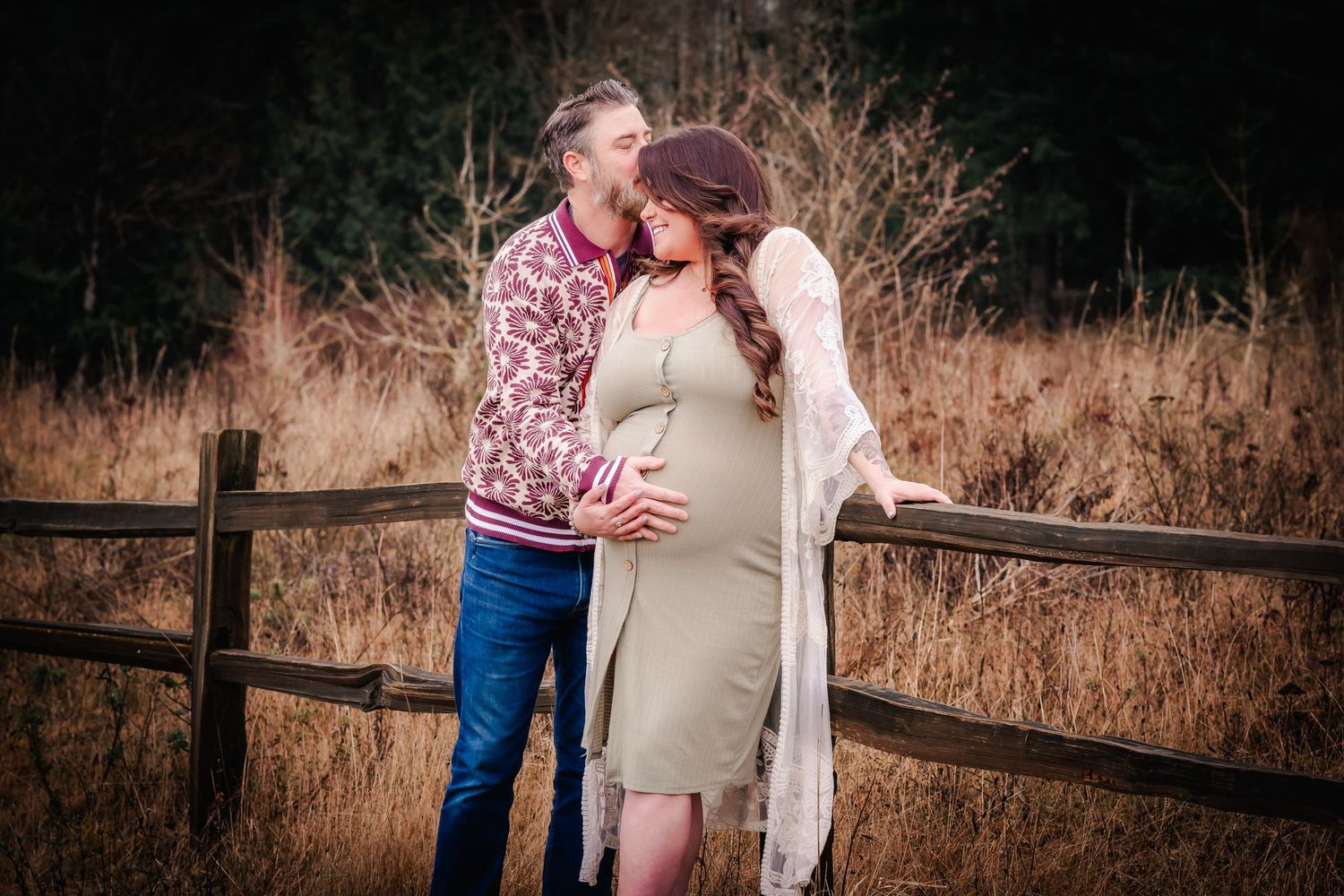 Couple sharing a tender moment by a wooden fence, woman in olive maternity dress with white cardigan, man in floral shirt.
