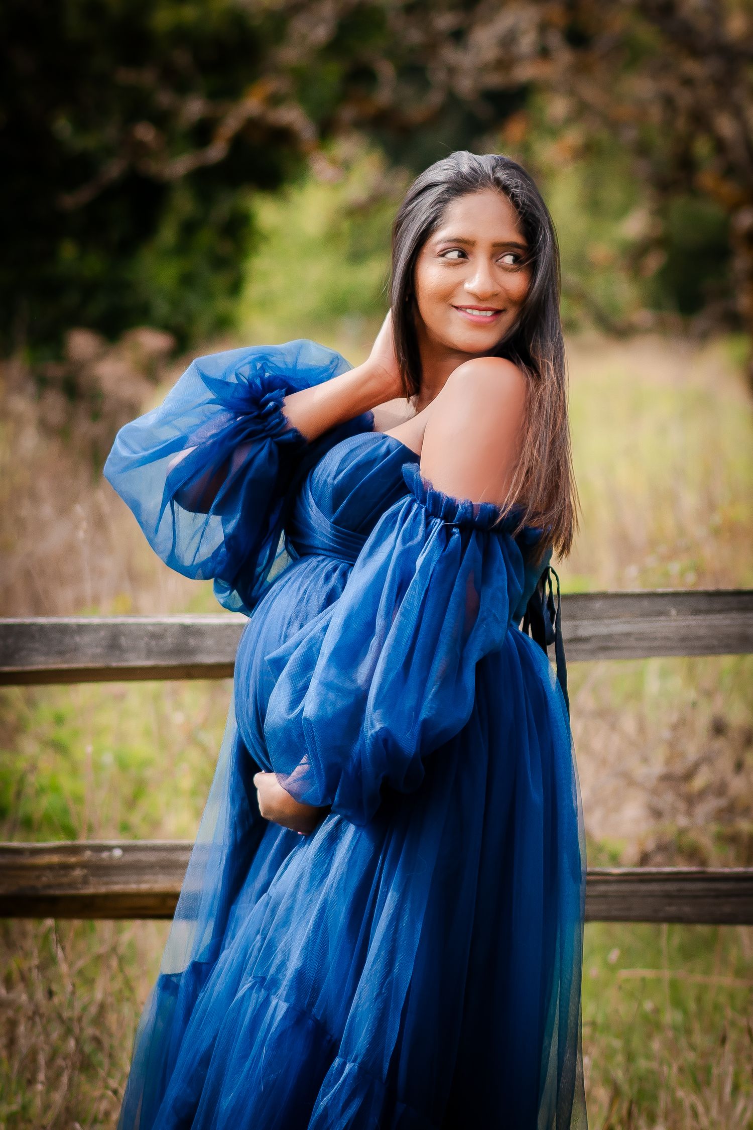 A person in a flowing royal blue dress poses against a wooden fence in a grassy field.