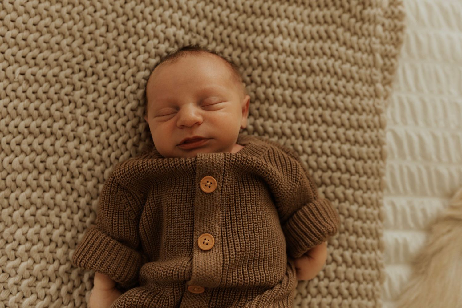 Sleeping newborn wearing brown knit cardigan rests on textured beige blanket.