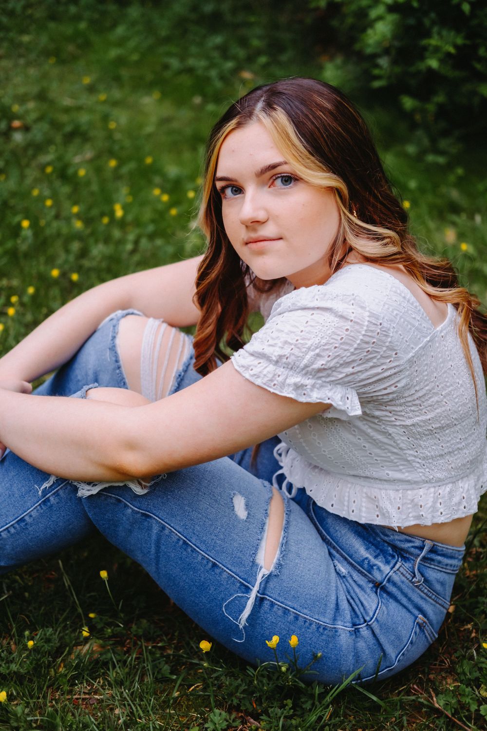Person in distressed jeans and white eyelet crop top sits on grass with yellow wildflowers in background.