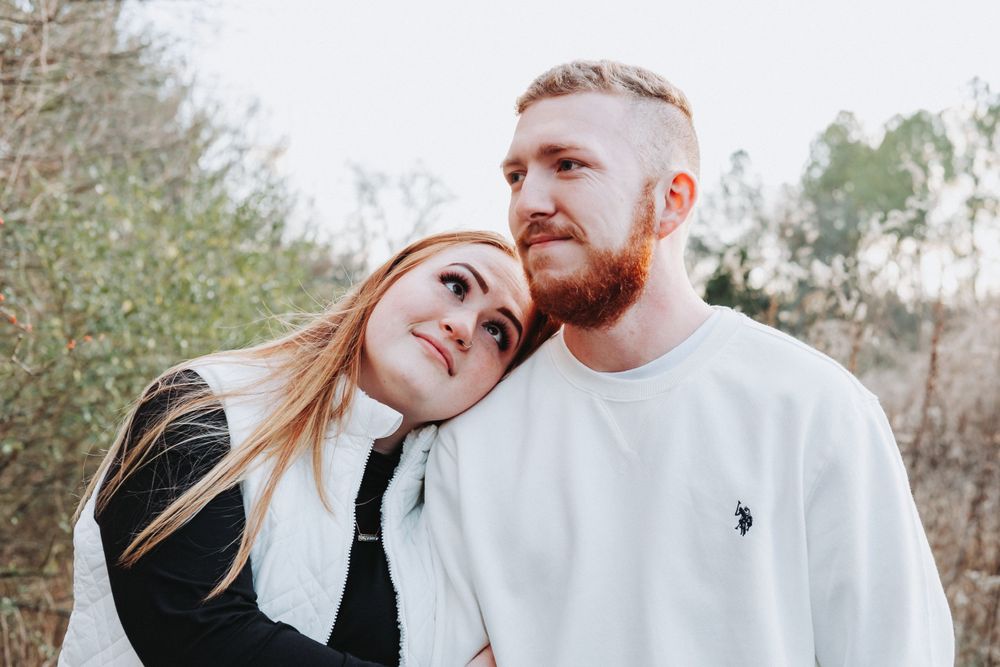 Couple poses together outdoors on a winter day with trees in background and wearing casual clothing.