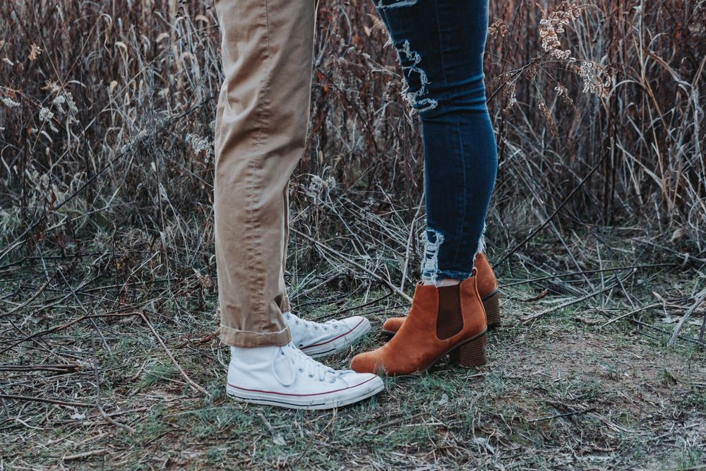 Two pairs of feet standing close together in brown boots and white sneakers against a backdrop of dried grass.