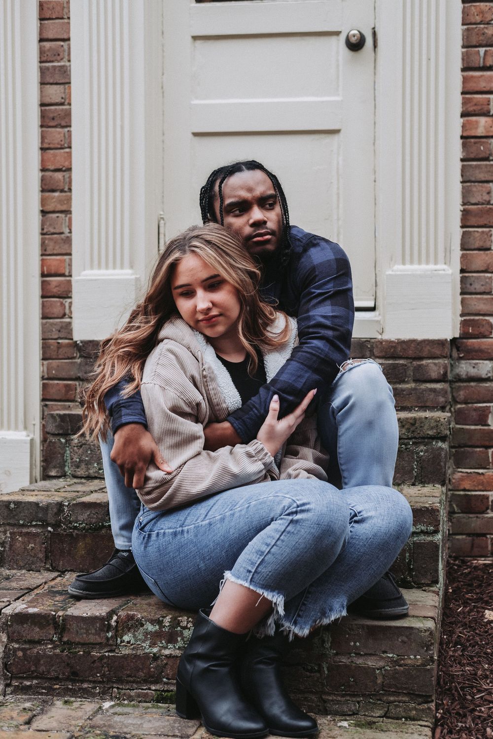 A couple sits close together on brick steps in front of a white door, wearing casual denim and cozy sweaters.