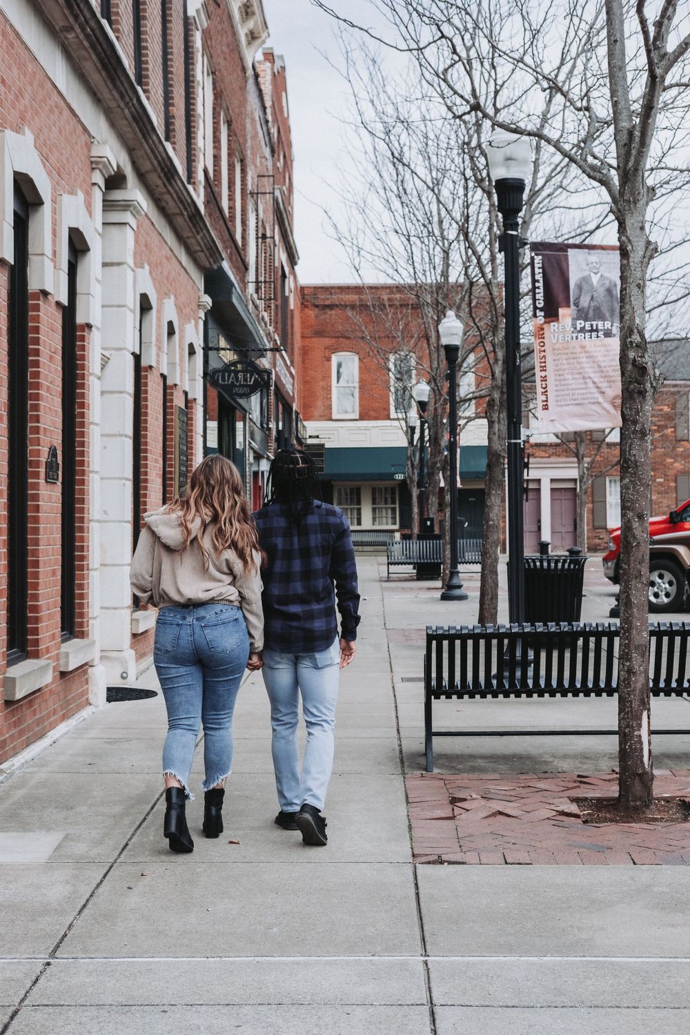 Two people stroll down a brick-lined small town sidewalk on a cool day with historic storefronts and street lamps visible.