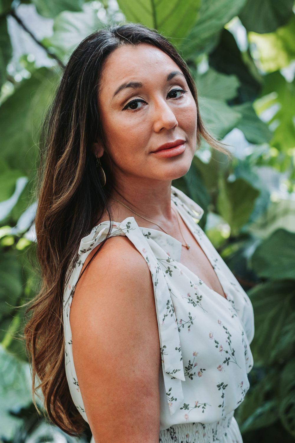 A person in a white floral dress poses outdoors against lush green foliage in soft natural lighting.