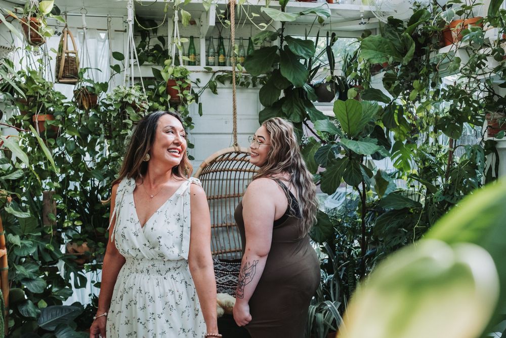 Two people share a joyful moment together in a lush greenhouse setting with hanging plants and natural light.