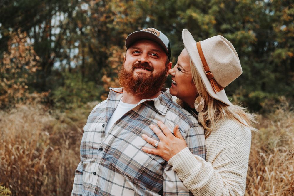 A couple embraces outdoors in autumn wearing casual attire with plaid flannel and knit sweater against a natural backdrop.