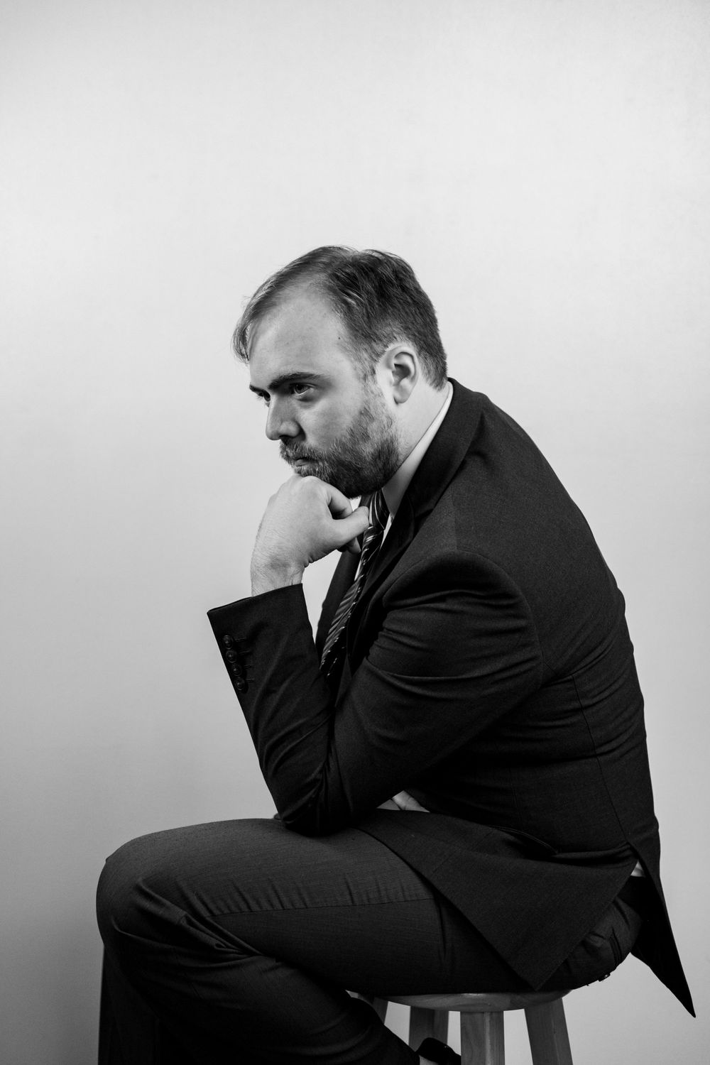 Black and white portrait of person in suit sitting on stool in contemplative pose against plain background.