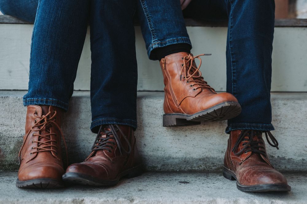 Two pairs of brown leather lace-up boots with dark blue jeans sitting on concrete steps.