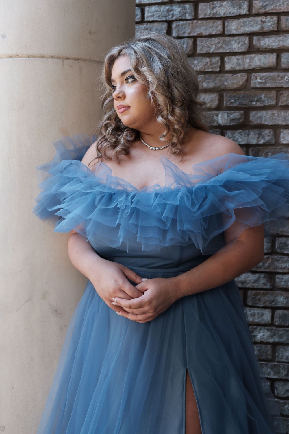 Romantic portrait in ethereal blue tulle gown with ruffled off-shoulder neckline against brick wall backdrop.