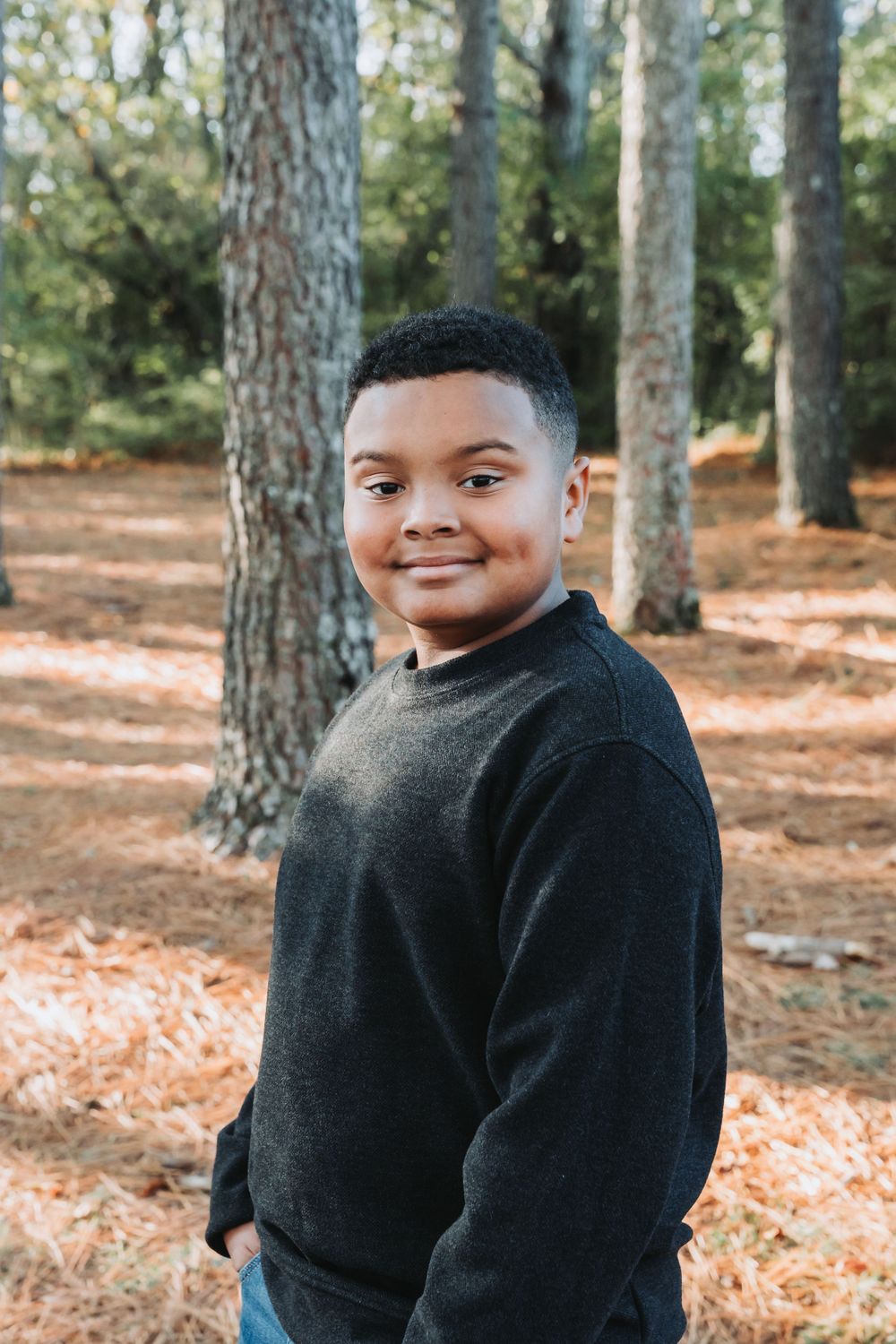 Young person in a black sweater poses outdoors in a wooded area with pine trees and fallen leaves in the background.