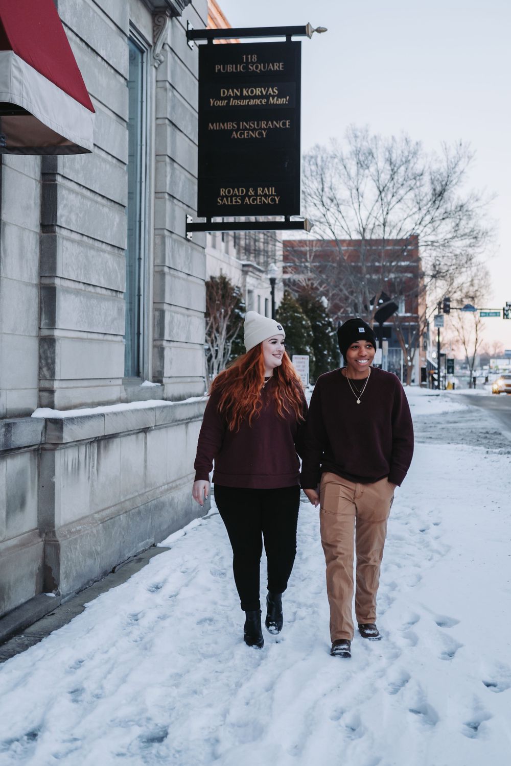 Two people walking together on a snowy winter sidewalk in front of a building with black signage.