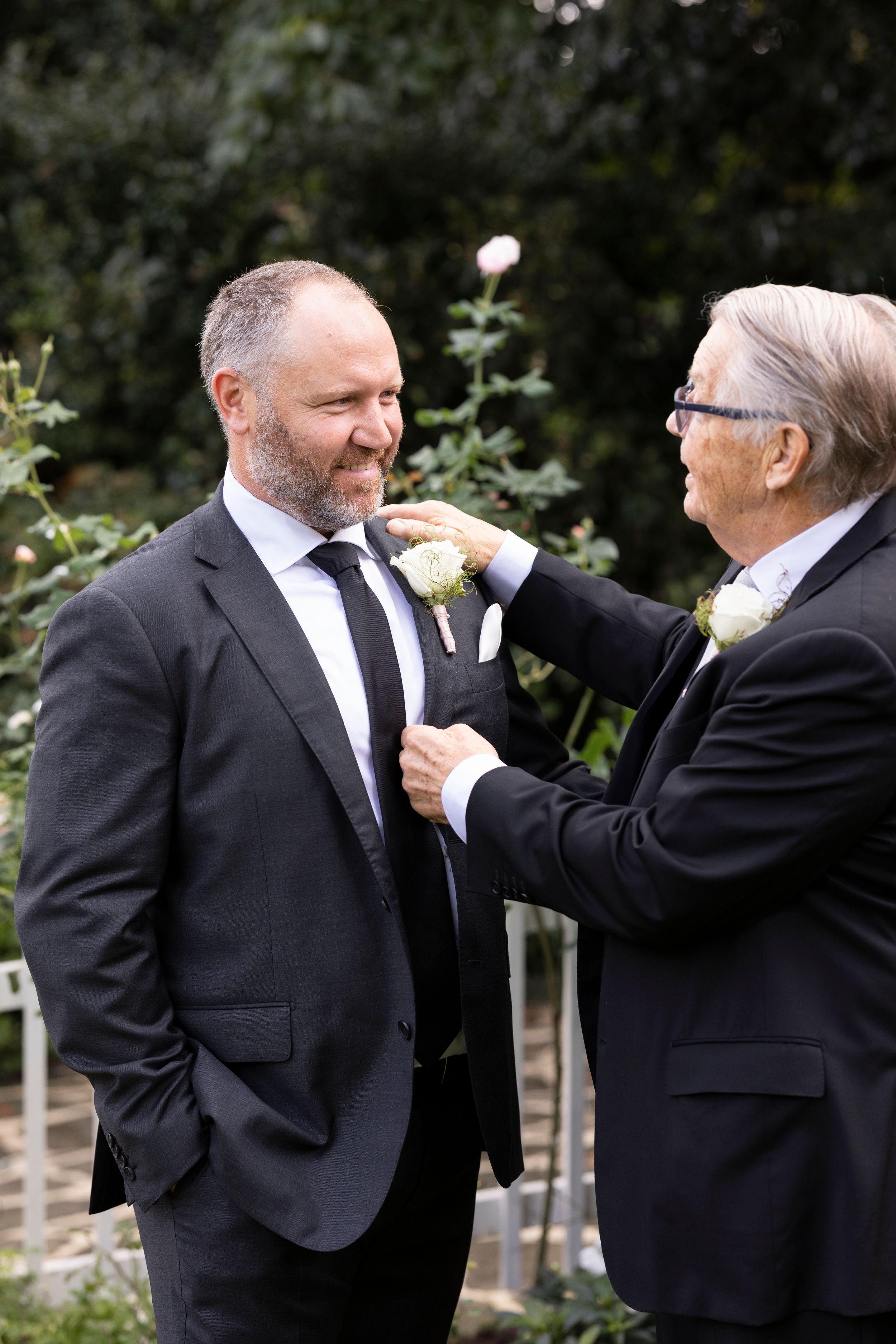 Two men in suits pinning boutonniere flower during wedding preparation.