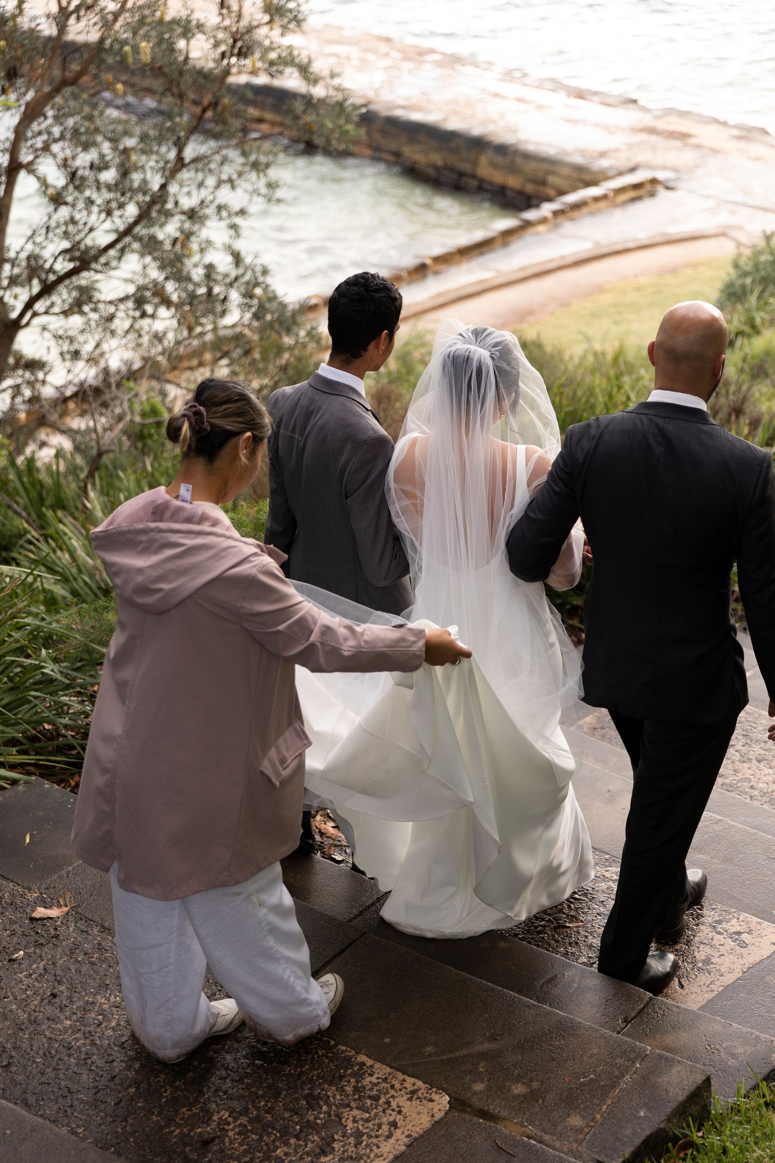 Wedding party helping with bride's dress while walking down coastal stone steps.