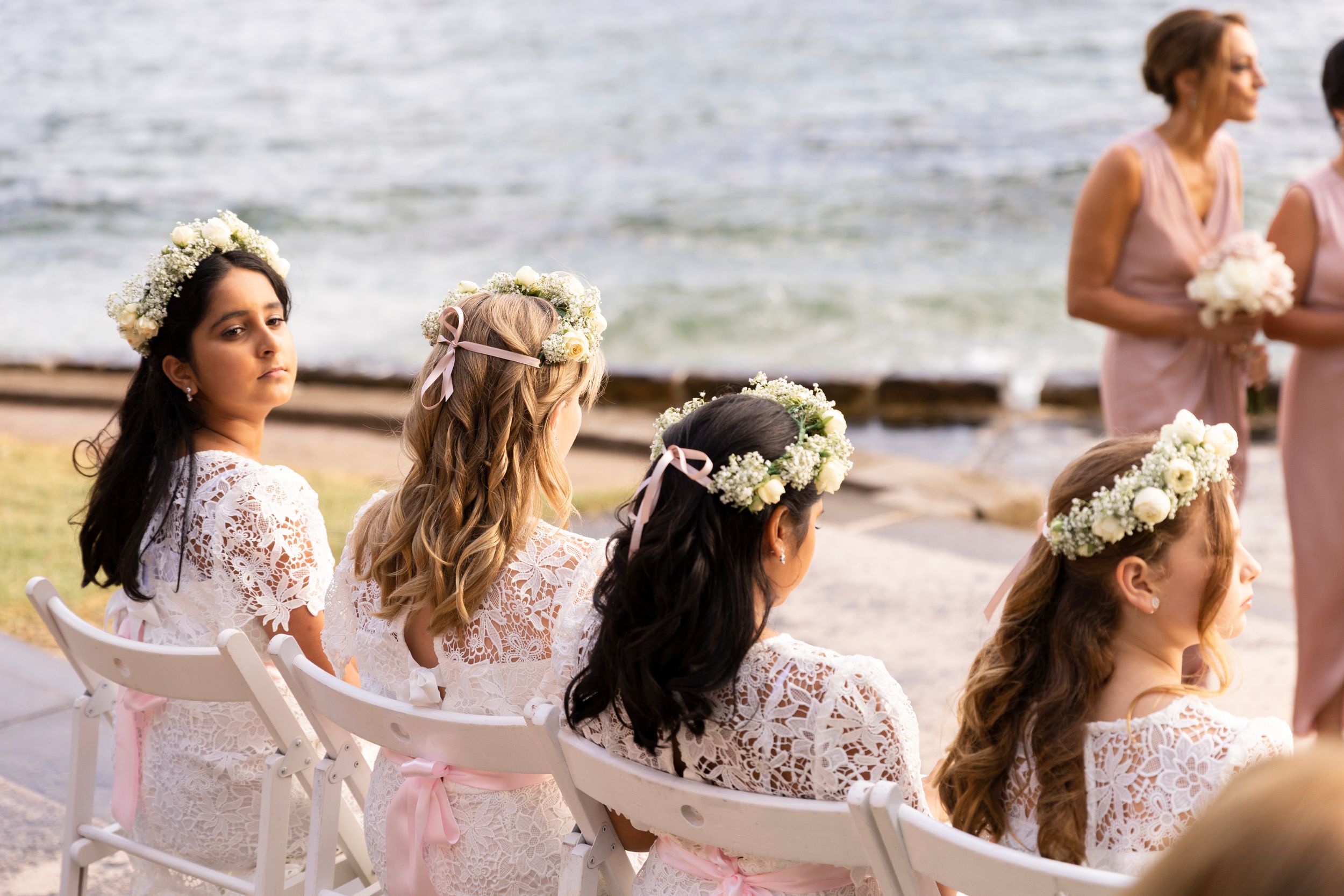 Row of bridesmaids in white lace dresses wearing floral crowns seated by the ocean.