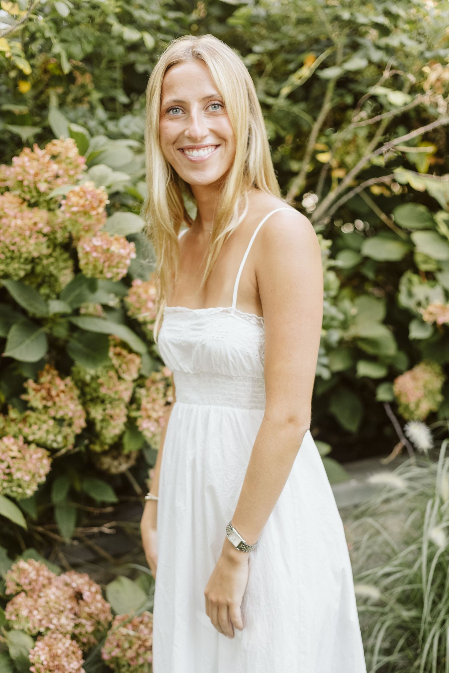 A woman in a white sundress stands in front of blooming hydrangea bushes.
