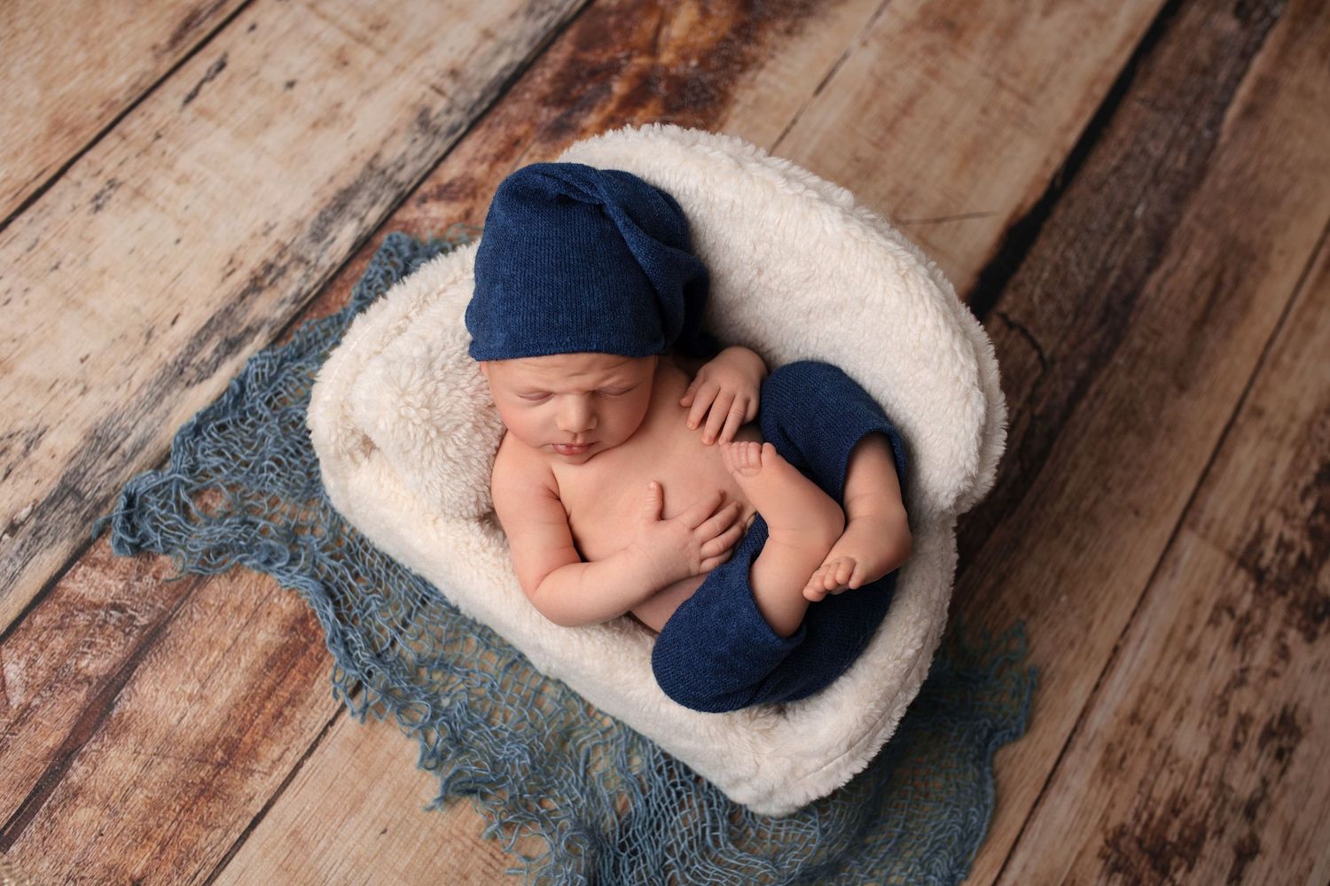 Sleeping newborn wearing navy blue accessories resting on white cushion against wooden floor.