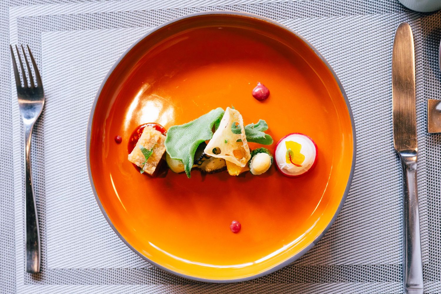 Elegantly plated gourmet dish on an orange ceramic plate with cutlery on a gray placemat.