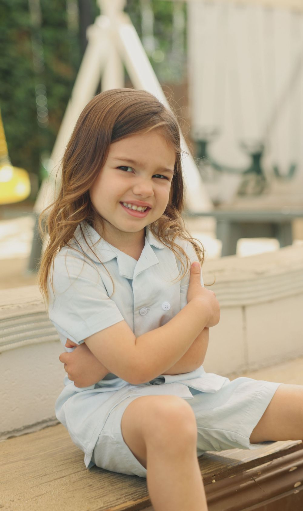 Smiling young girl in light blue dress sitting with arms crossed at playground.