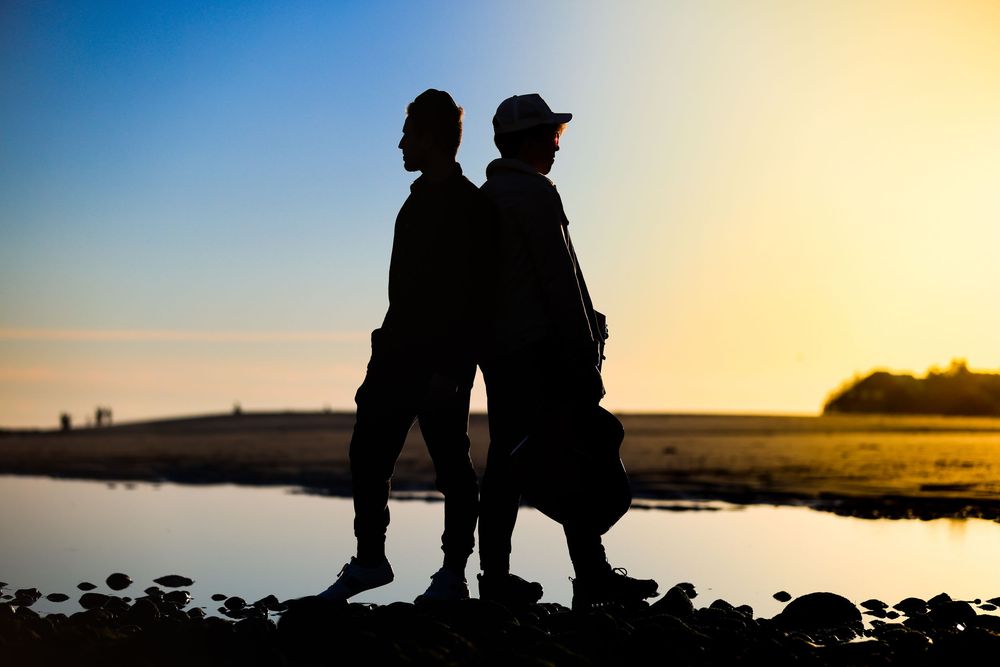 Two people silhouetted back-to-back on a rocky beach during a serene golden sunset.