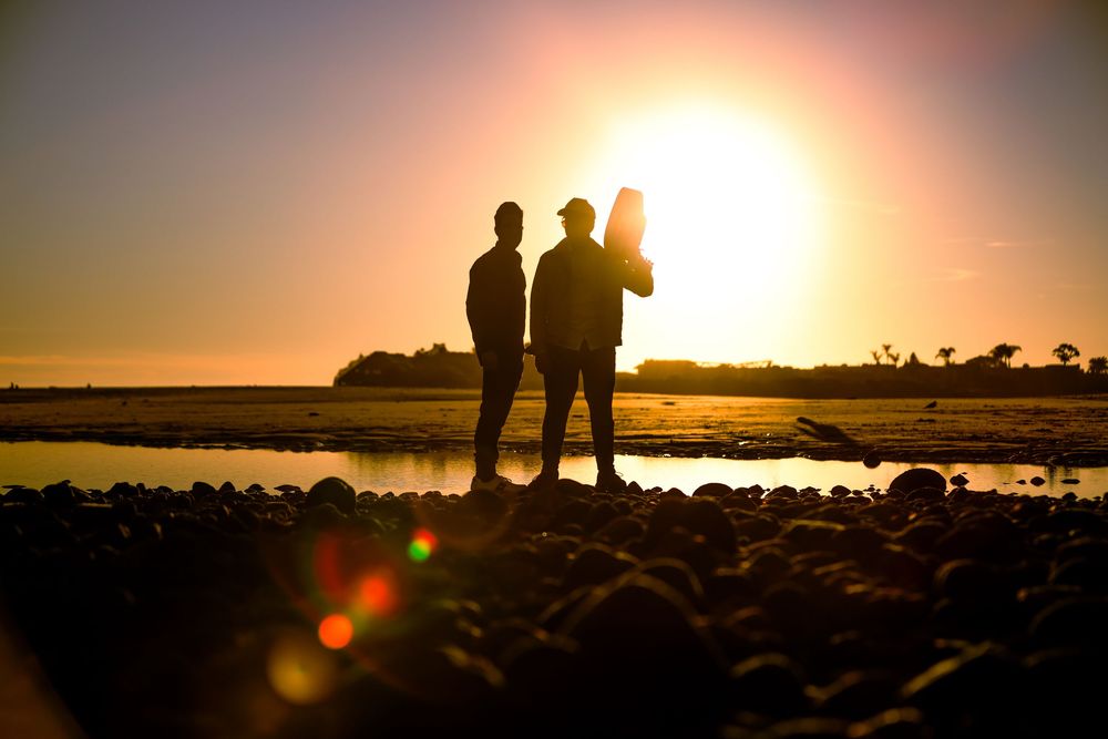 Three people standing in silhouette on a rocky shore with a bright golden sunset behind them.