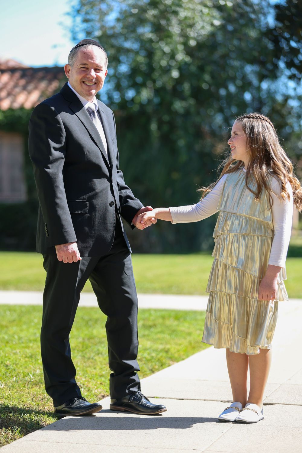 Man in black suit holding hands with young girl in gold dress outdoors.