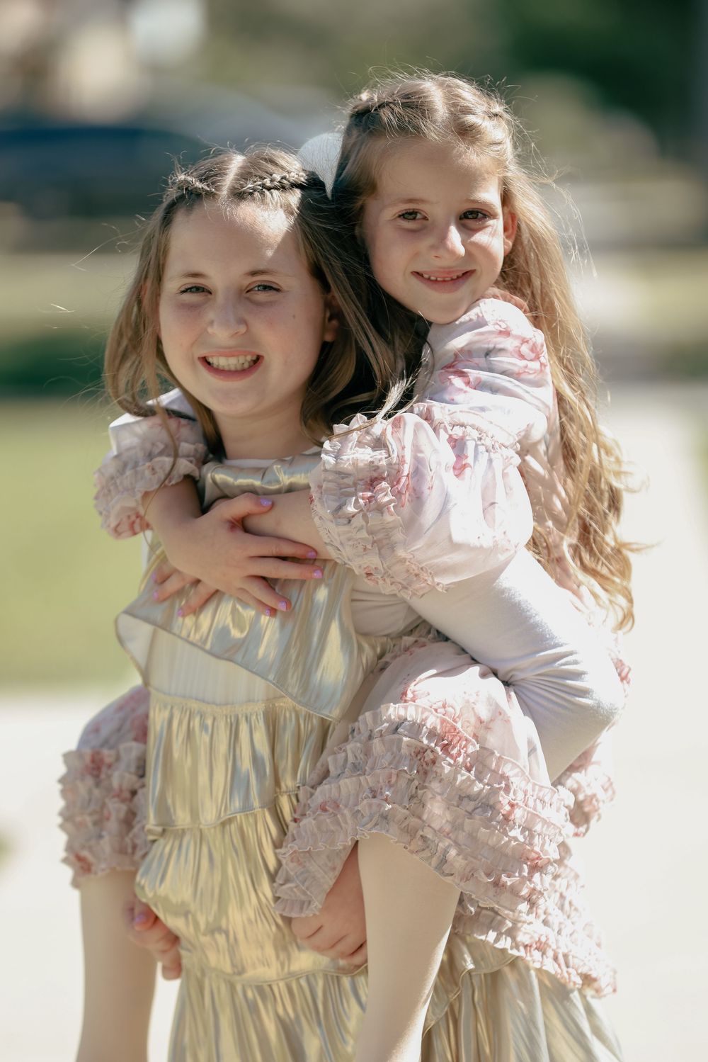 Two young girls in formal dresses hugging and smiling outdoors in sunlight.