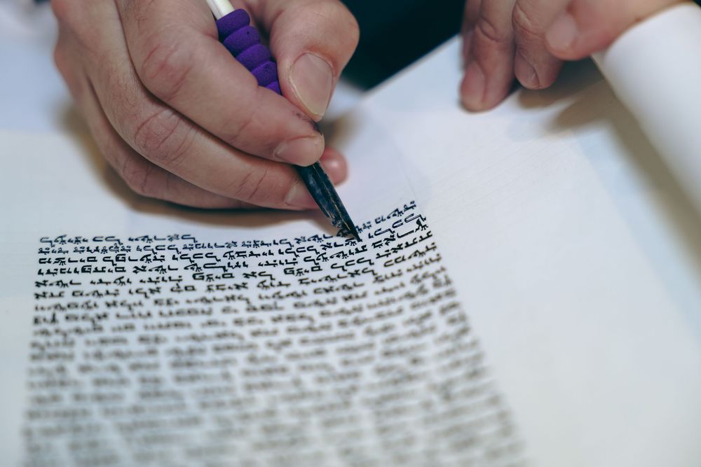 Hands writing Hebrew text on a Jewish religious scroll with a pen.