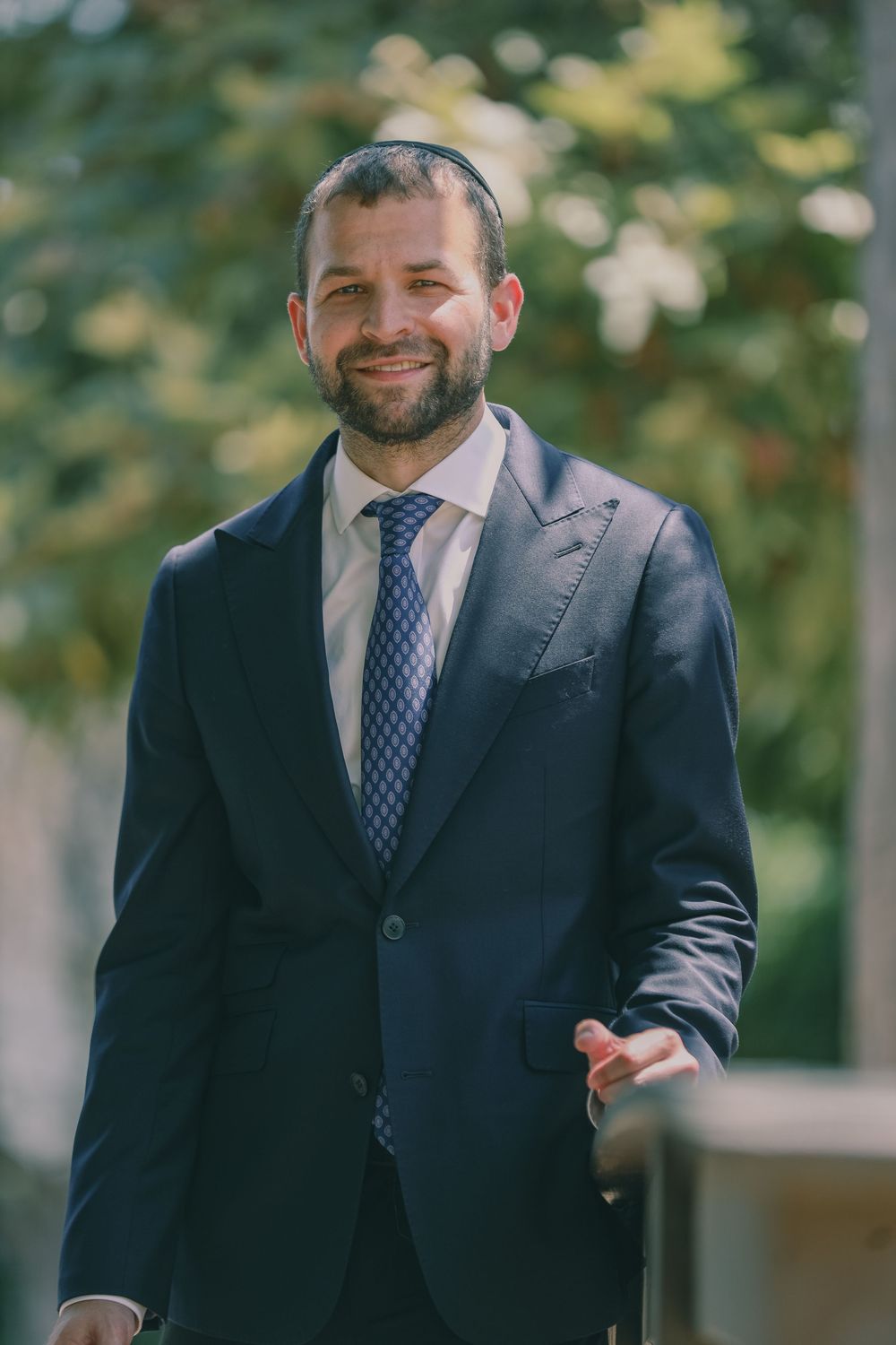 Smiling bearded man in navy suit and kippah outdoors in natural setting.