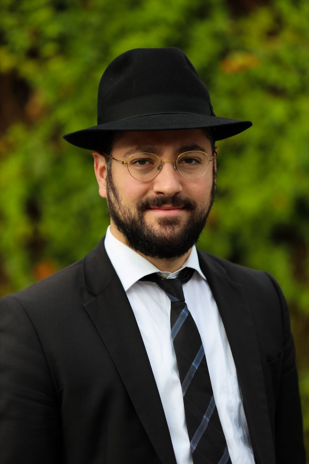 Bearded man in black suit, fedora hat, and glasses posing outdoors.