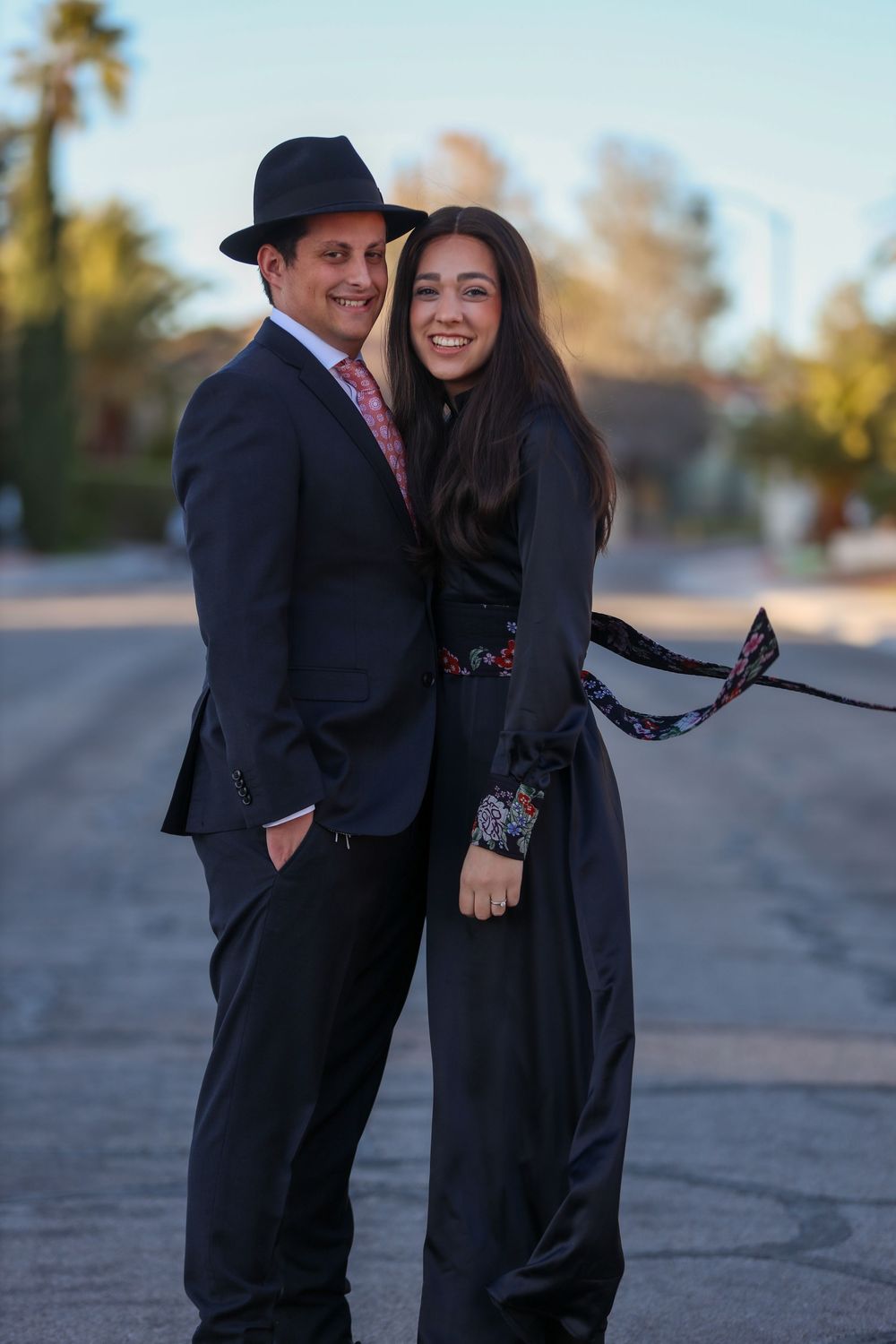 Couple in formal black outfits posing on a suburban street at dusk.