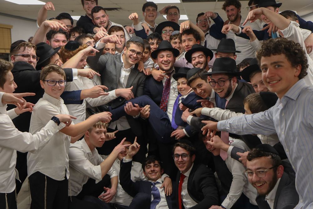 Large group of young men in formal attire celebrating together indoors, lifting one person up.