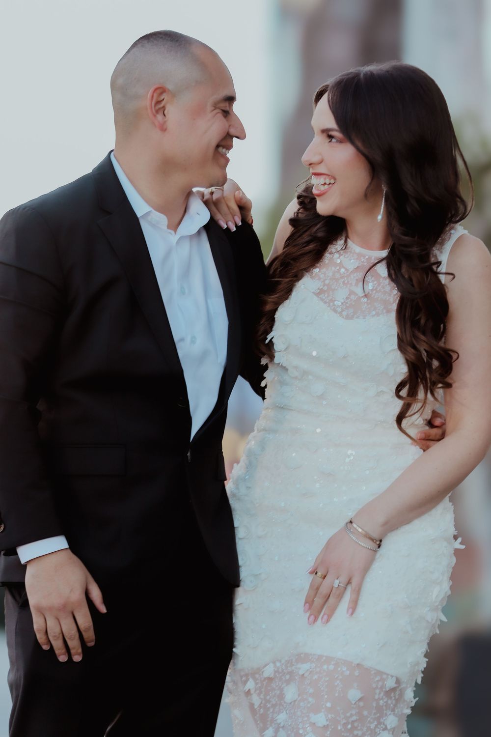 Smiling bride in white lace dress and groom in black suit at outdoor ceremony.